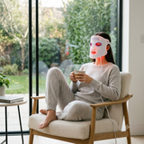 Person sitting in a chair wearing a LED face mask and holding a phone, with a garden view outside.